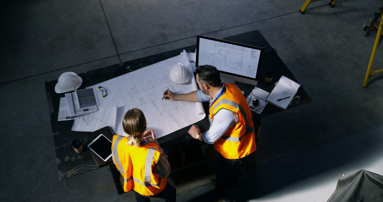 Two people in orange vests looking at blueprints spread out on a desk