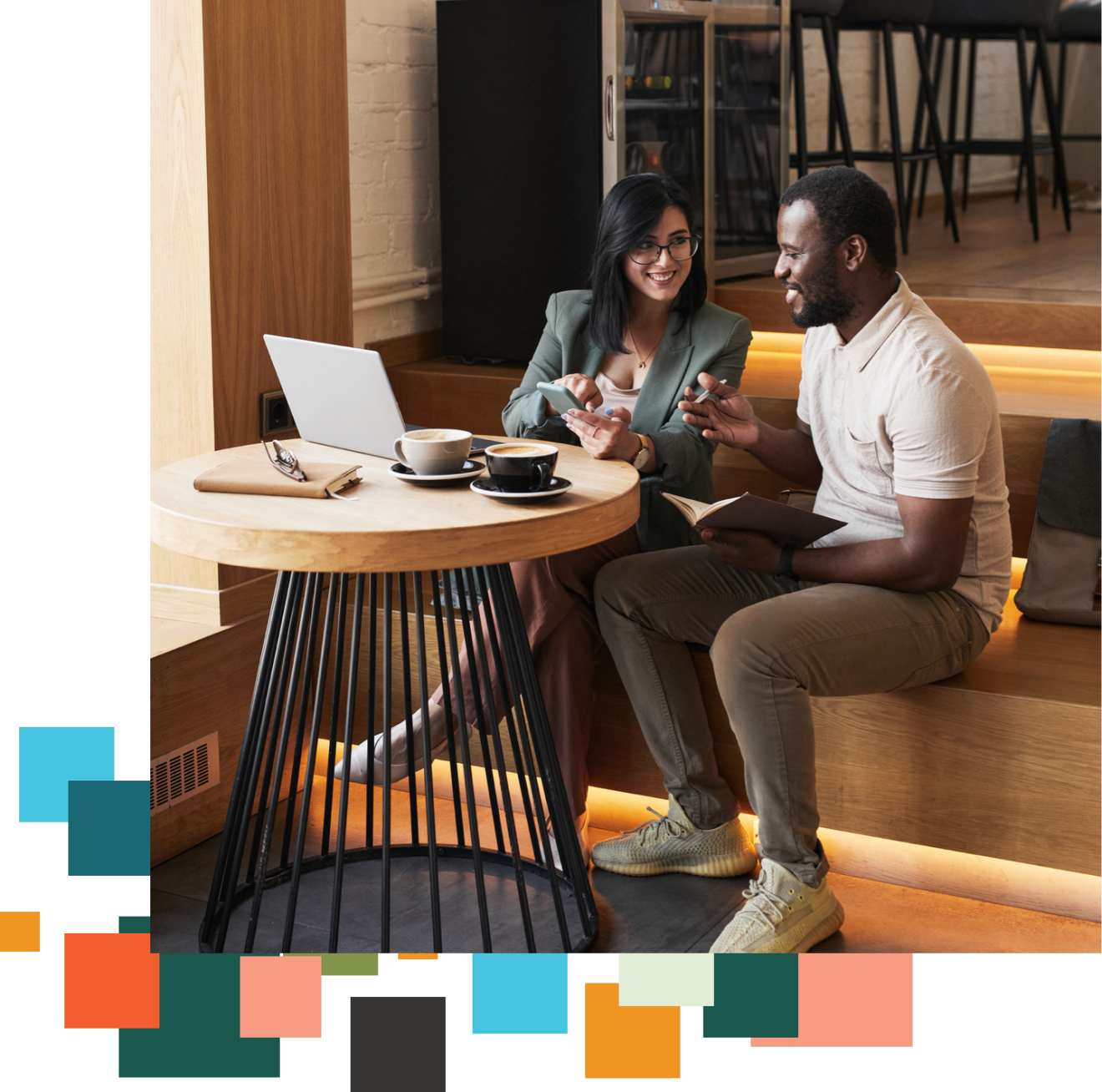 Two people sit at a table together and talk while looking at a laptop